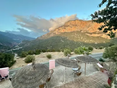 Traditional thatched umbrellas on stone terrace overlooking dramatic mountain landscape in Al Oued, Morocco, with golden hour lighting illuminating rocky peaks and winding valley roads below