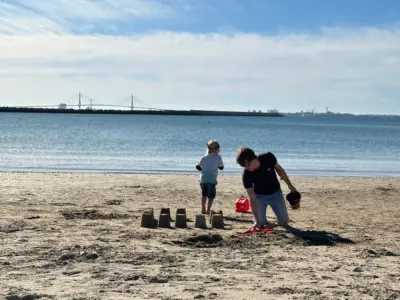 Father and child building sandcastles on sandy beach in El Puerto de Santa María, Spain, with blue water and distant bridge visible under cloudy sky